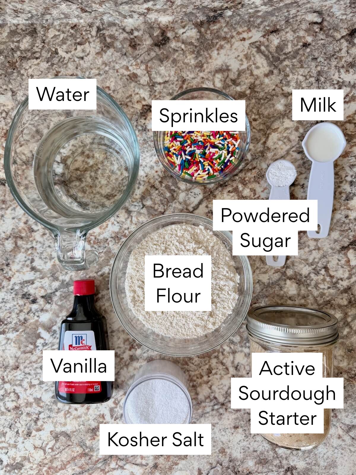The ingredients to make funfetti sourdough focaccia. Each ingredient is labeled with text. They include water, sprinkles, bread flour, vanilla, kosher salt, active sourdough starter, powdered sugar, and milk.