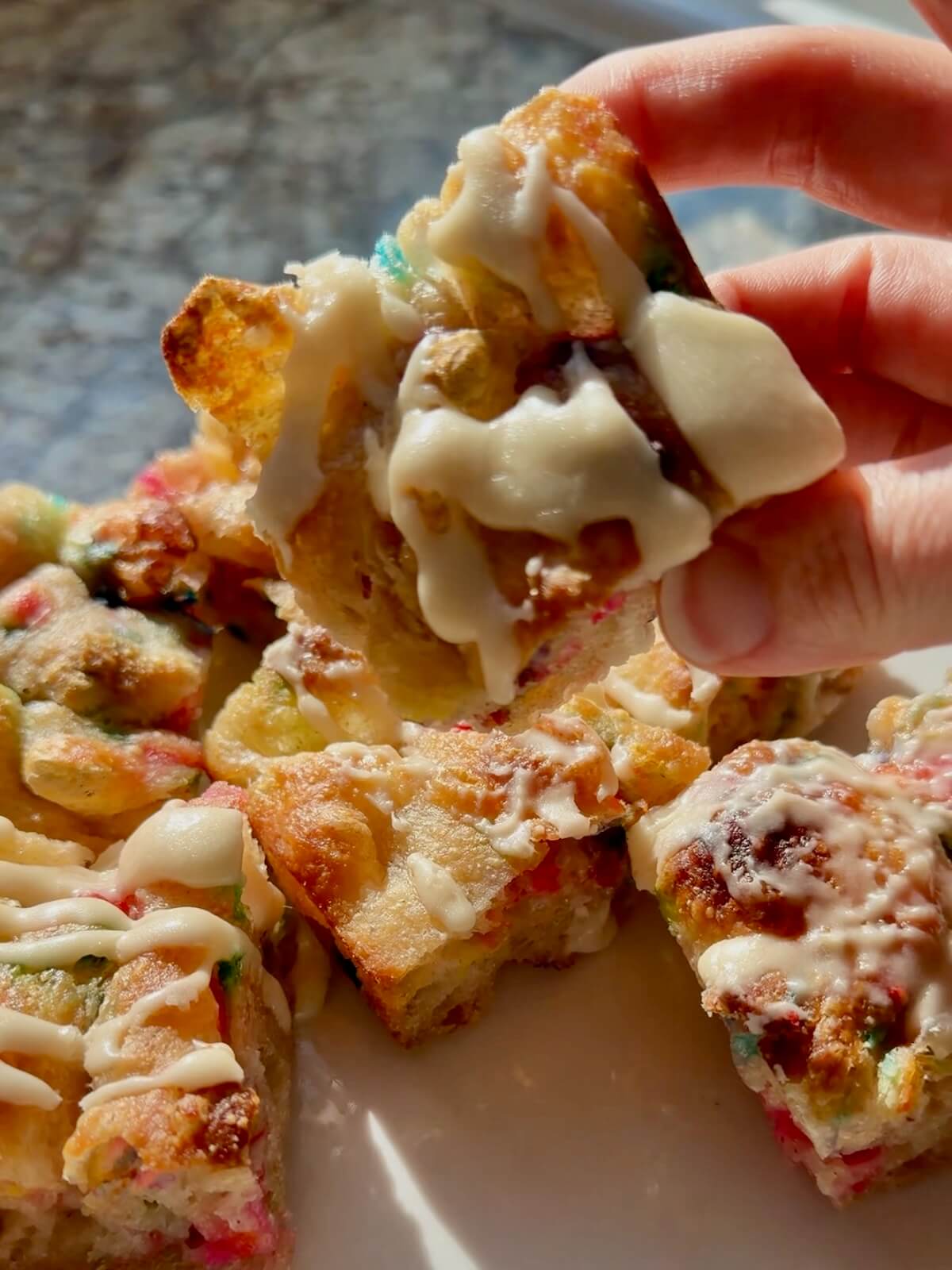 A plate of funfetti sourdough focaccia pieces drizzled with icing. A hand is holding up one piece to the camera.