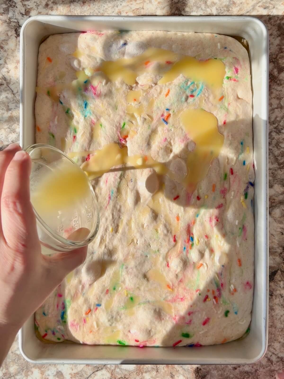 Melted butter being poured on top of a proofed funfetti sourdough focaccia dough in a baking pan.