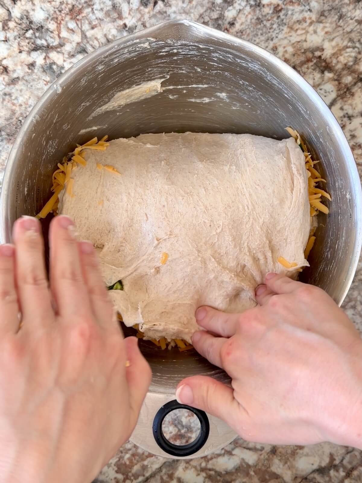 Two hands stretching and folding diced jalapeƱos and shredded cheddar cheese into sourdough bread dough.