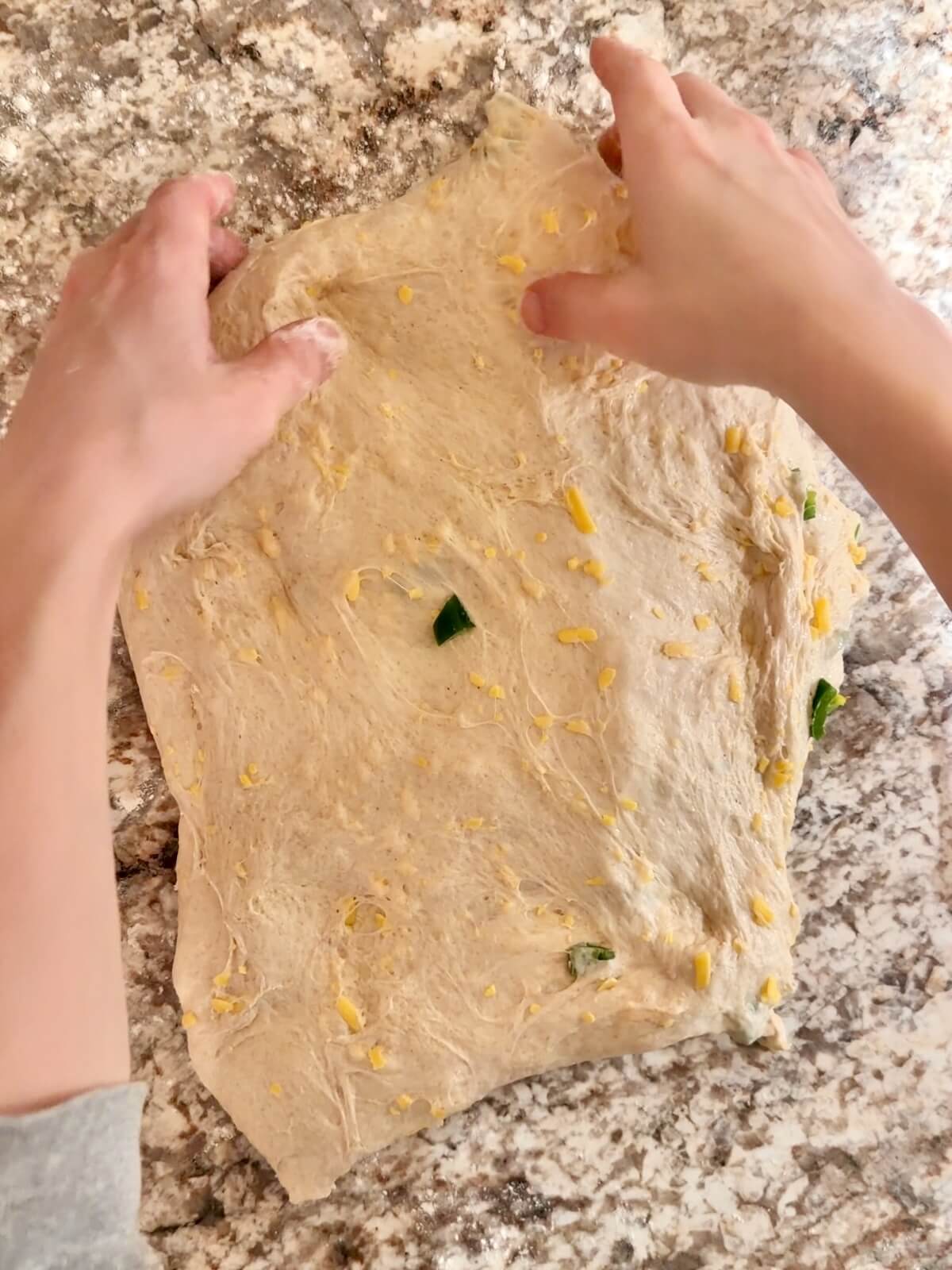 Two hands stretching jalapeƱo cheddar sourdough bread dough into a rectangle on a floured counter.