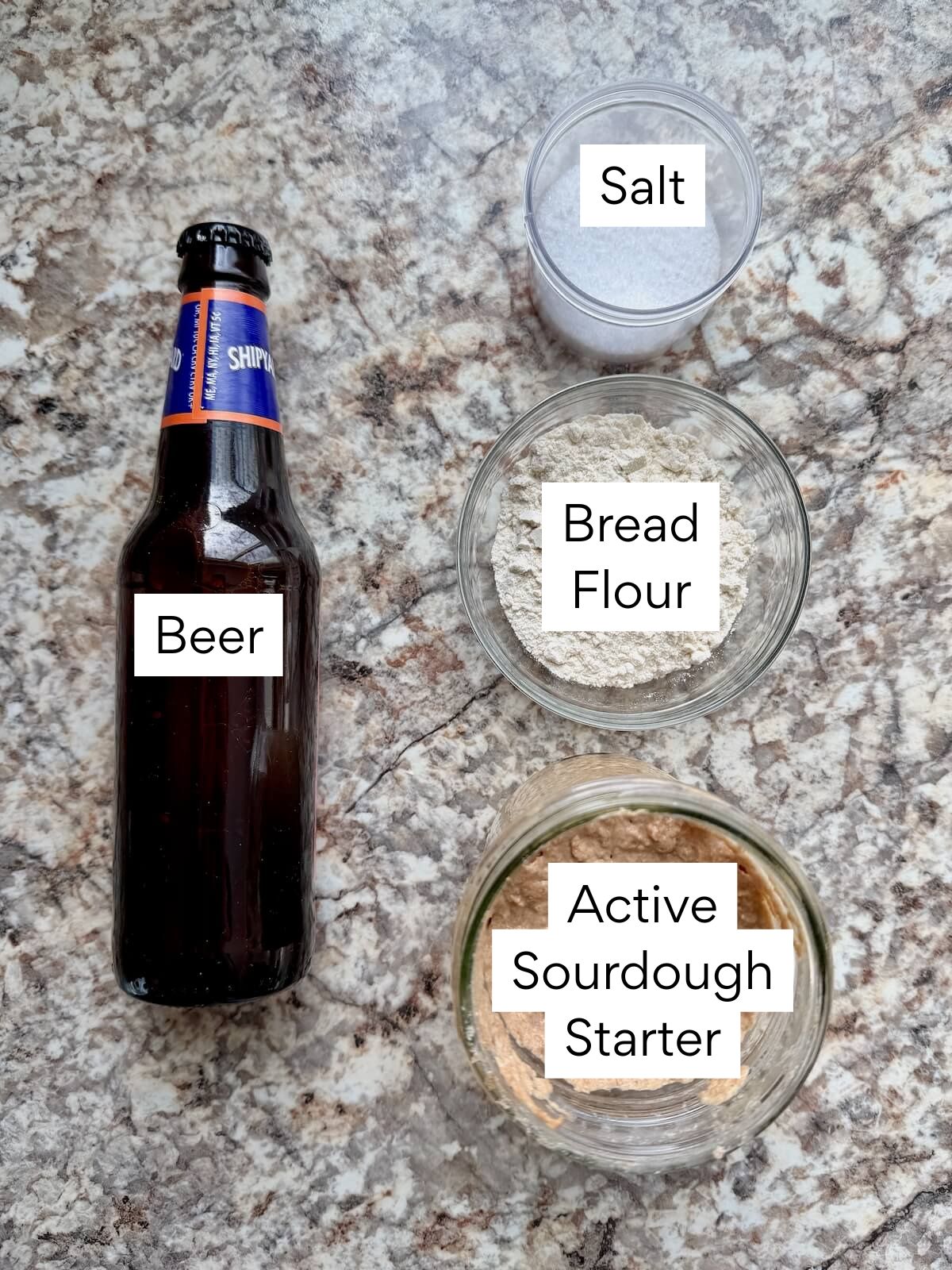 The ingredients to make beer sourdough bread. Each ingredient is labeled with text. They include beer, salt, bread flour, and active sourdough starter.