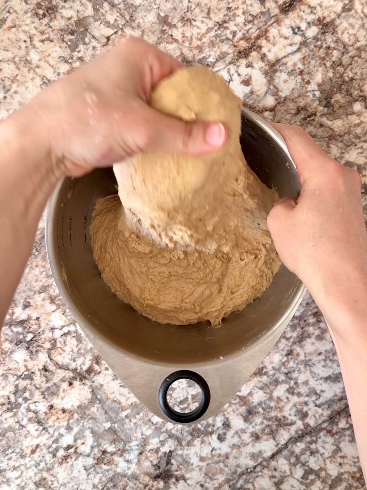 Two hands stretching the dough up and out of the mixing bowl.