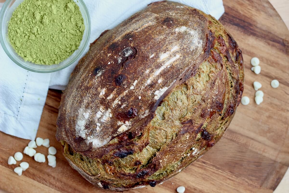 A whole loaf of matcha sourdough bread on a wooden cutting board. Around it are scattered white chocolate chips and a small bowl of matcha powder.