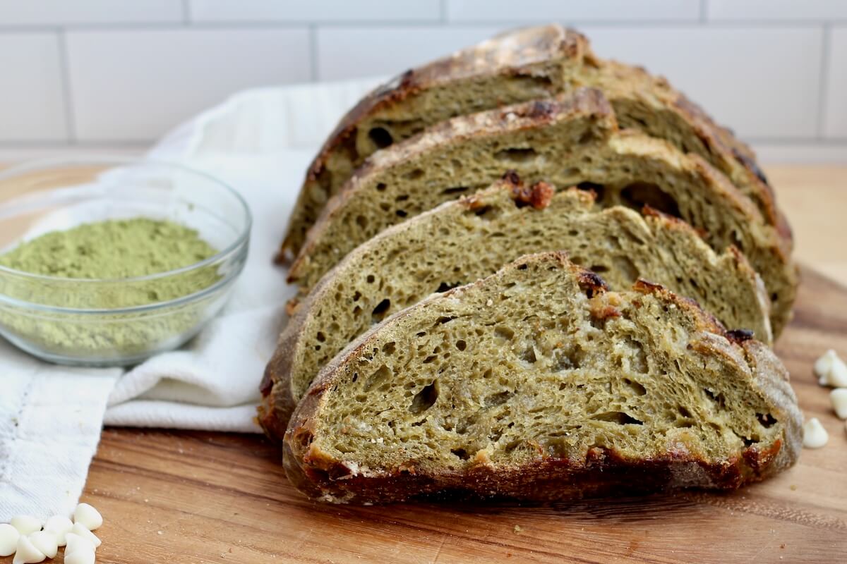 Slices of white chocolate matcha sourdough bread on a wooden cutting board. There is a small bowl of matcha powder to the left of the bread and a few white chocolate chips to the right.