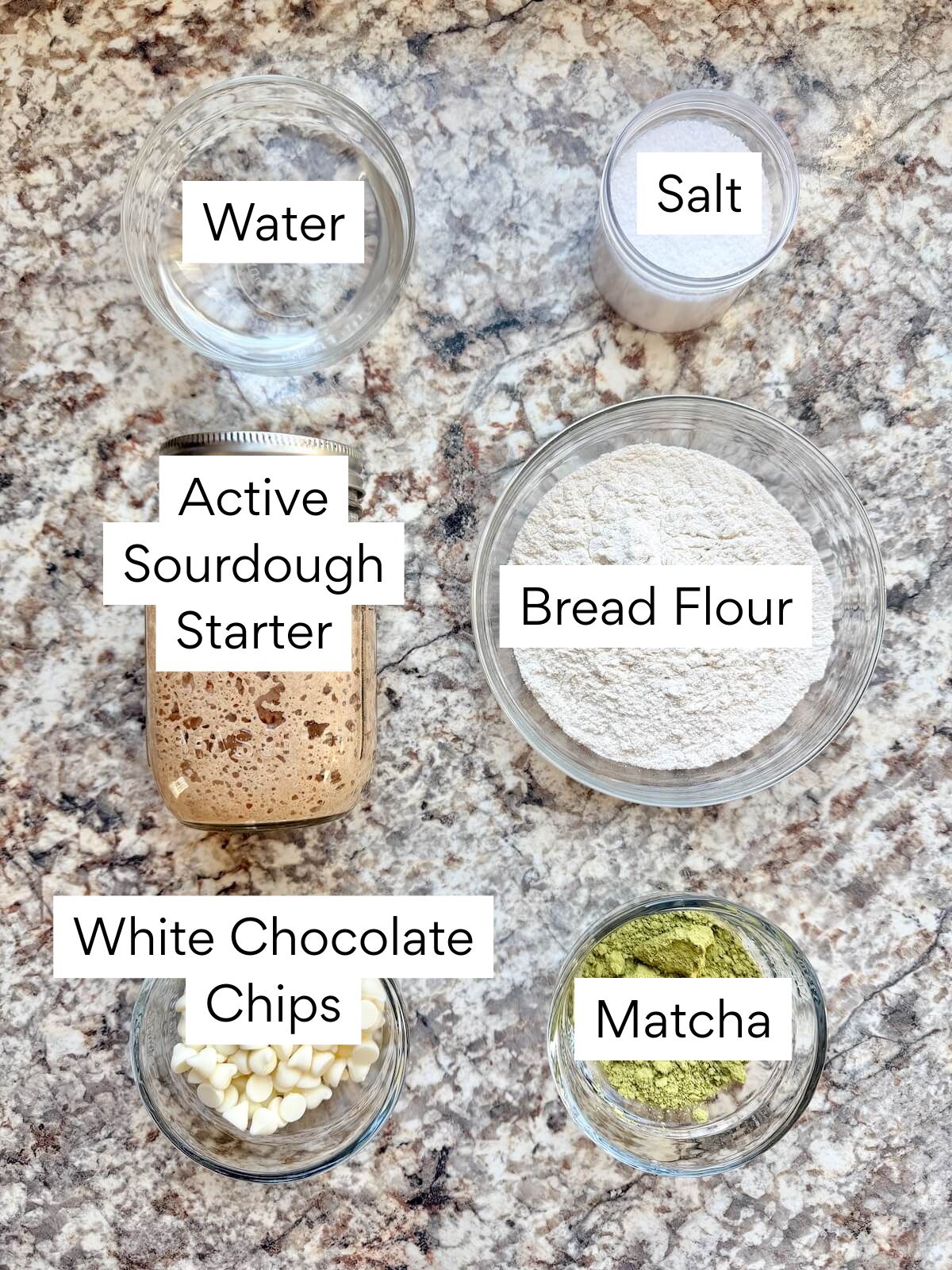 The ingredients to make white chocolate matcha sourdough bread. Each ingredient is labeled with text. They include water, active sourdough starter, white chocolate chips, salt, bread flour, and matcha.
