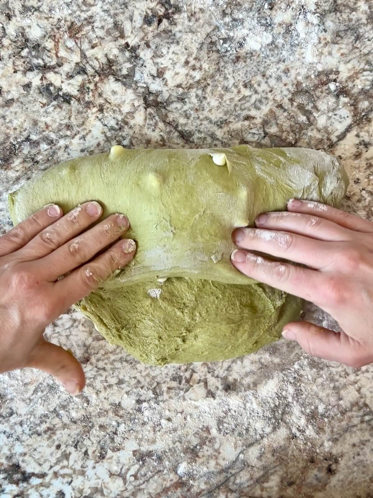 Two floured hands shaping matcha sourdough bread dough on a counter.