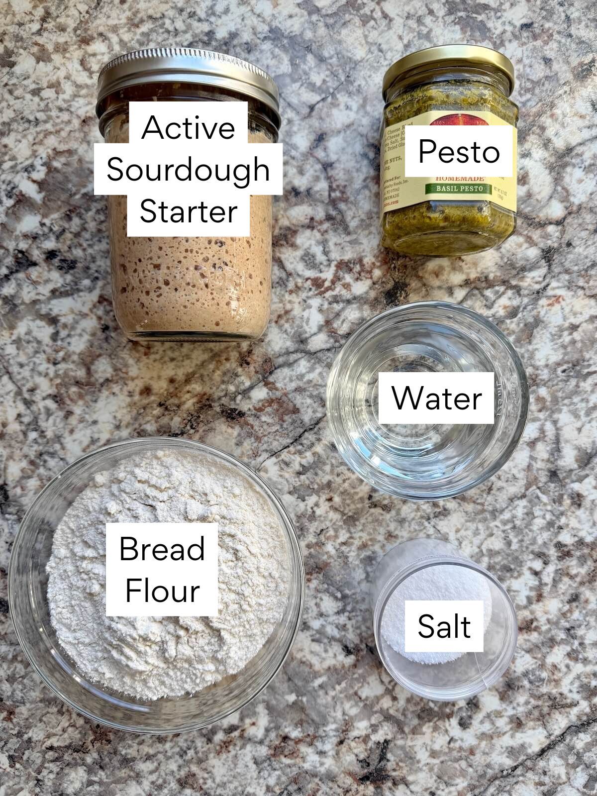 The ingredients to make pesto sourdough bread. Each ingredient is labeled with text. They include active sourdough starter, bread flour, pesto, water, and salt.