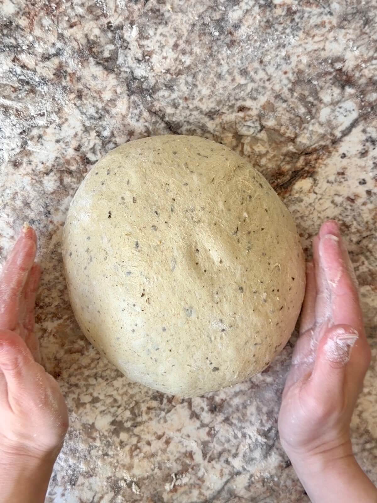 Pesto sourdough bread dough being shaped into a ball on a floured counter.