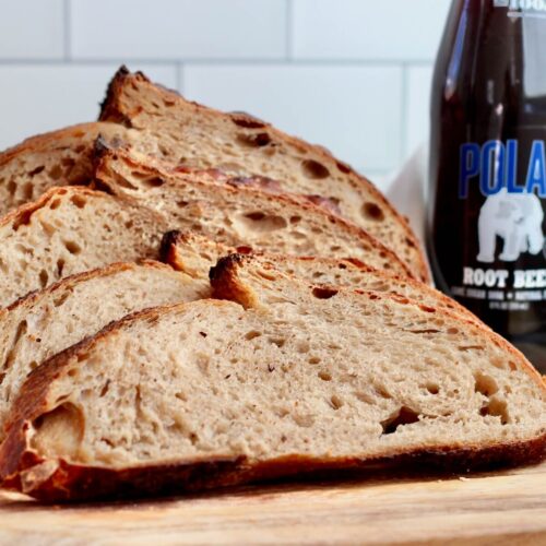 Slices of root beer sourdough bread on a wooden cutting board next to a bottle of root beer.