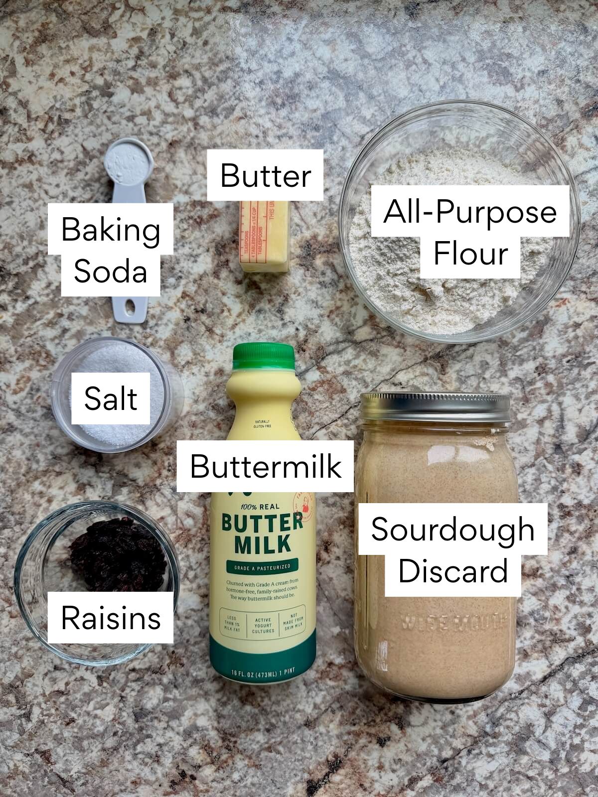 The ingredients to make sourdough Irish soda bread. Each ingredient is labeled with text. They include baking soda, butter, all-purpose flour, salt, buttermilk, sourdough discard, and raisins.