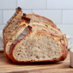 A sliced loaf of high protein sourdough bread on a wooden cutting board. There is a scoop of whey protein next to the loaf.