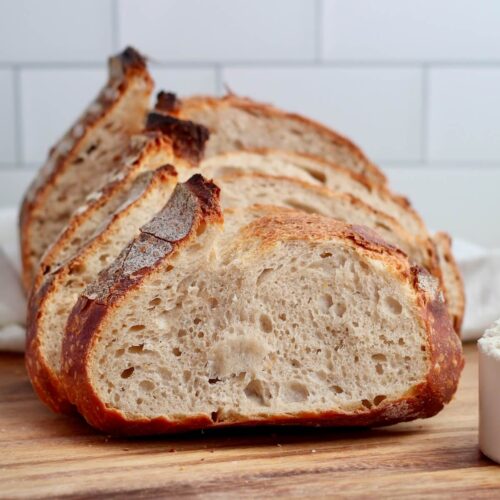 A sliced loaf of high protein sourdough bread on a wooden cutting board. There is a scoop of whey protein next to the loaf.