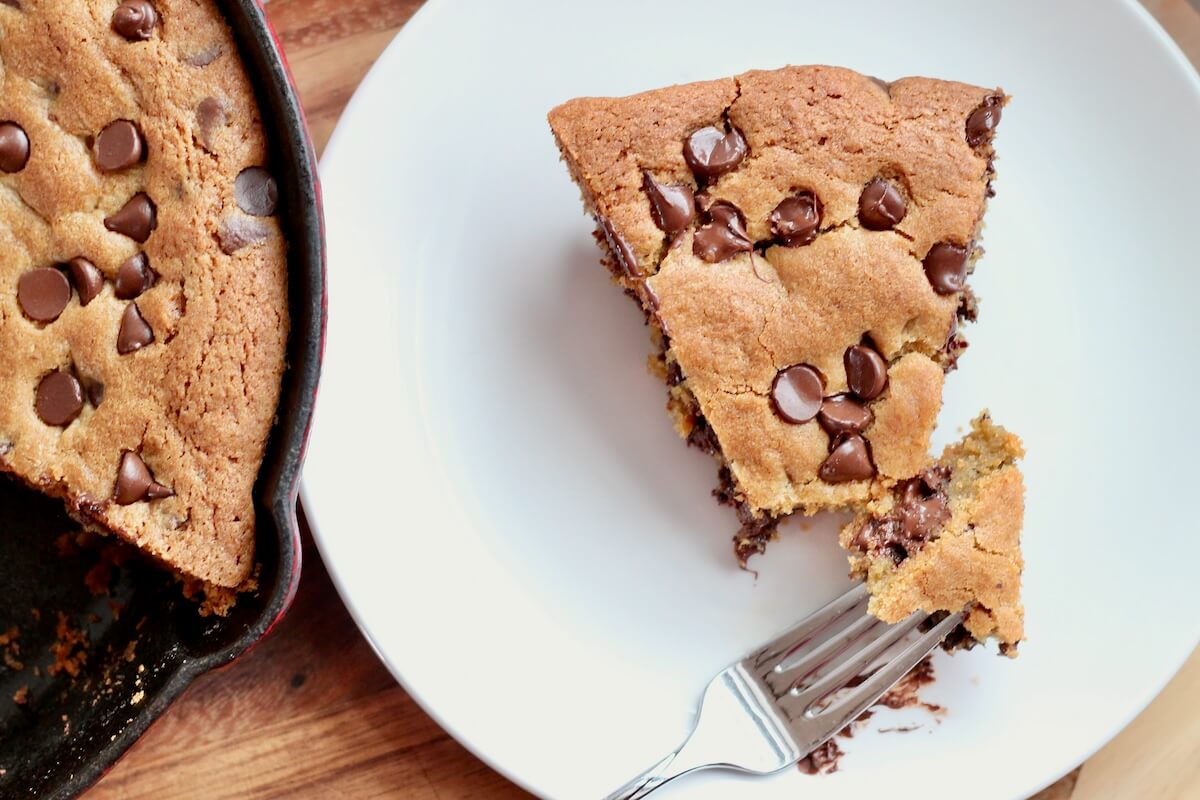 A slice of sourdough skillet cookie on a small plate. A fork took a small piece from the cookie and is resting on the plate.
