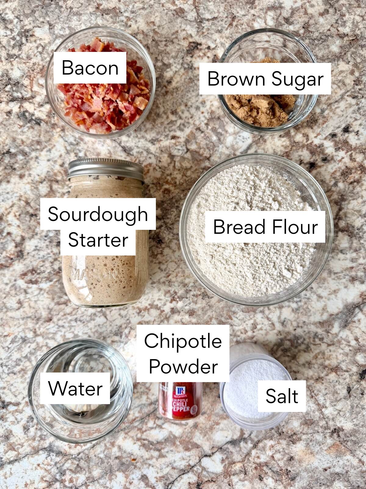The ingredients to make smoky brown sugar bacon sourdough bread. Each ingredient is labeled with text. They include bacon, brown sugar, sourdough starter, bread flour, water, chipotle powder, and salt.