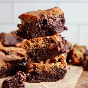 A stack of three sourdough brookies on a wooden cutting board with more brookies out of focus in the background.