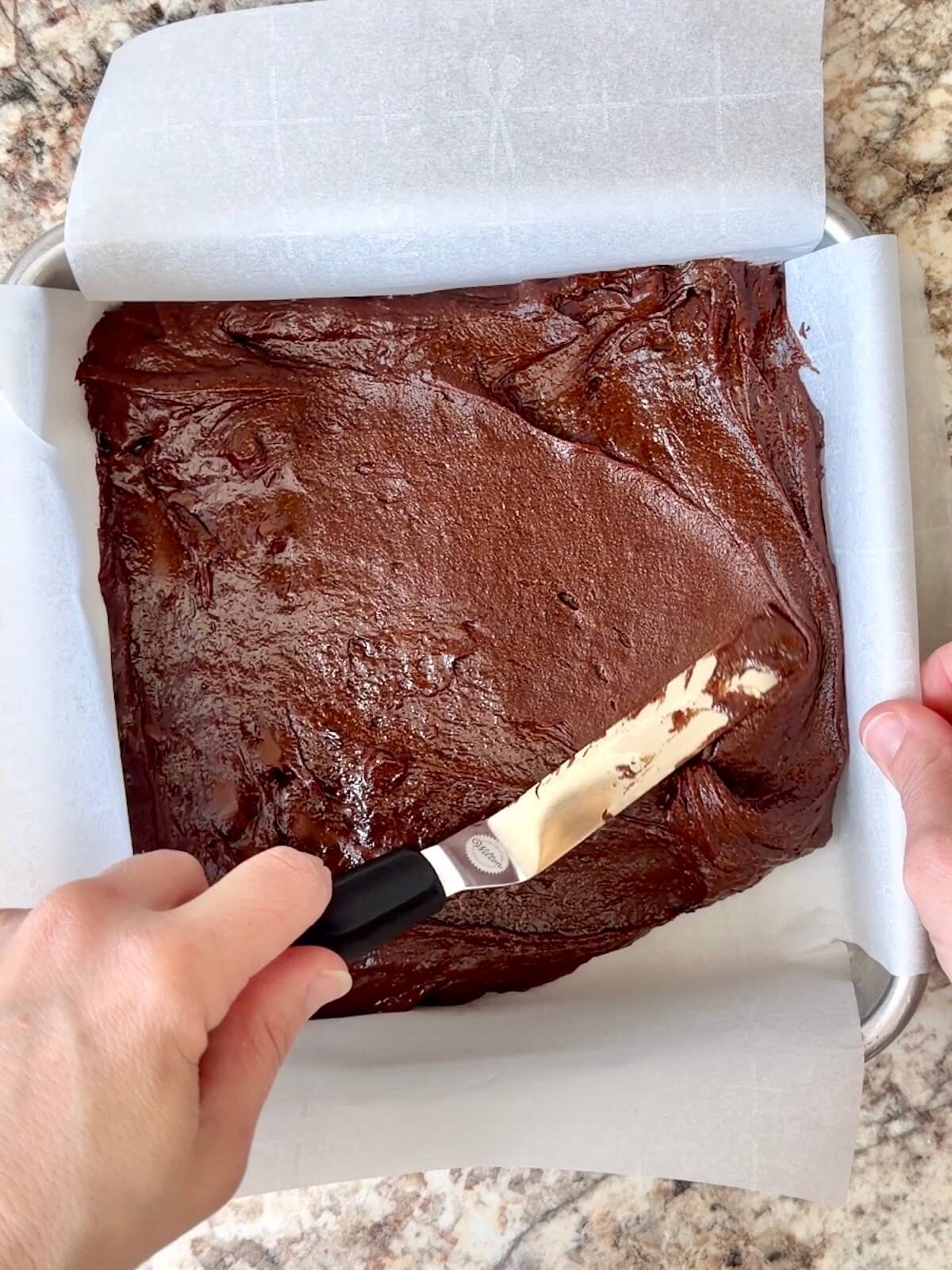 Sourdough brownie batter being spread into a parchment-lined, square aluminum pan.