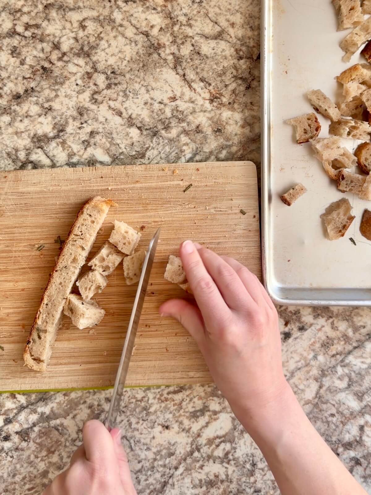 Two hands cubing sourdough bread on a cutting board with a baking sheet to the right.