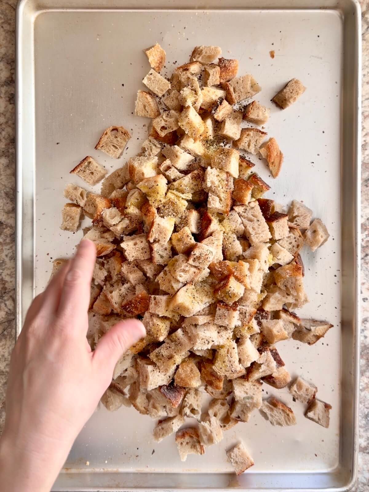 A hand seasoning the cubed sourdough bread on a baking sheet.