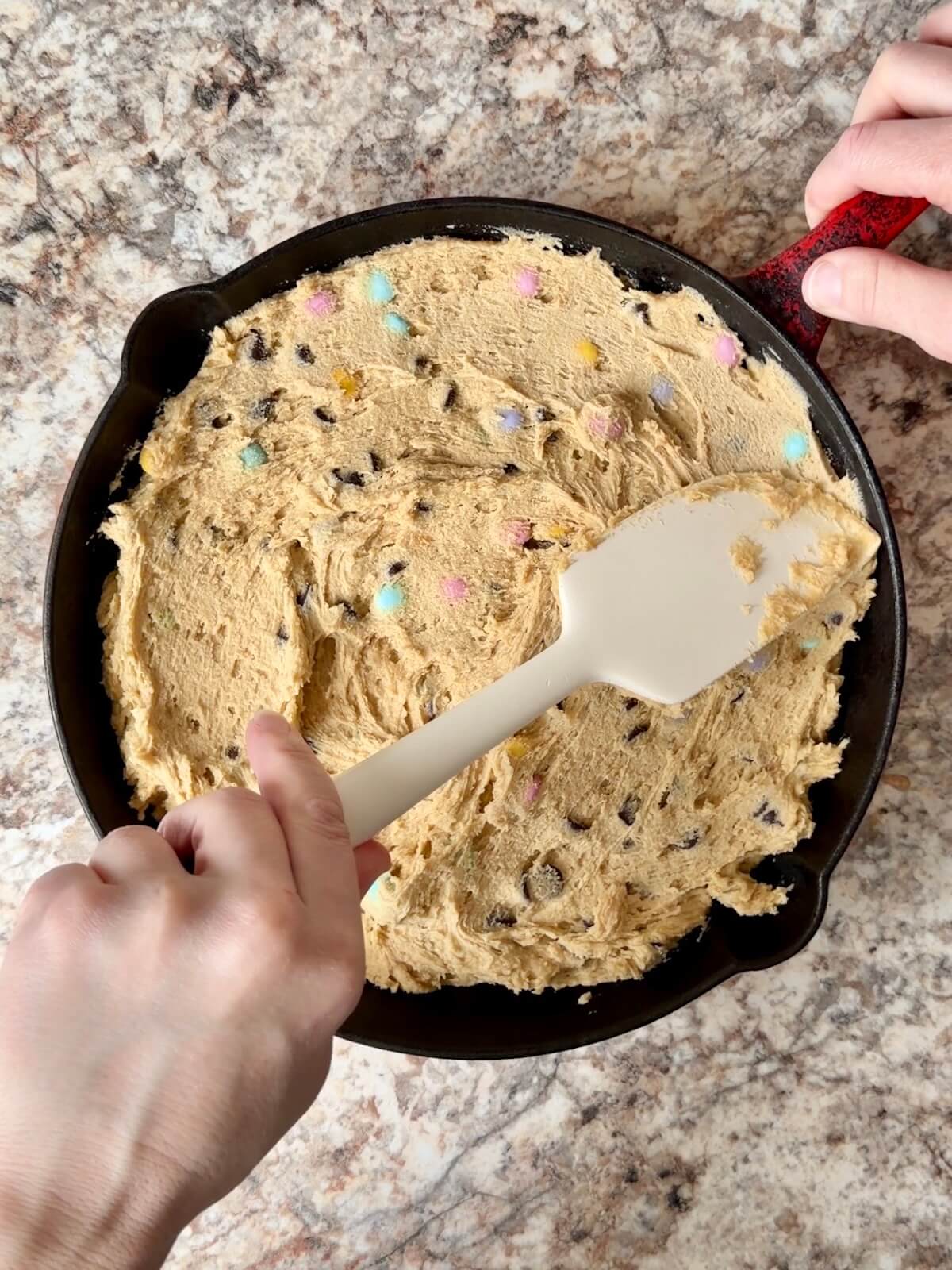 Two hands using a rubber spatula to spread sourdough discard cookie dough in a cast iron skillet.