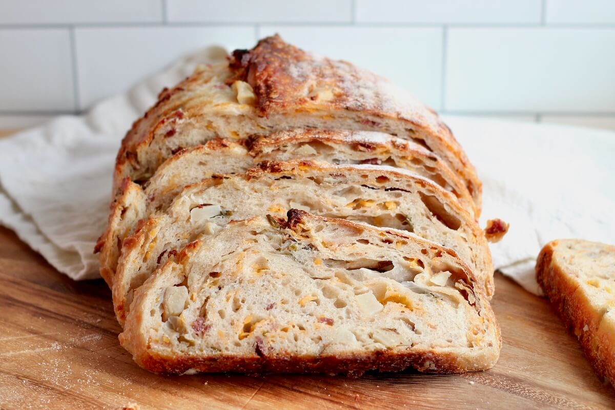 A sliced loaf of loaded baked potato sourdough bread on a wooden cutting board.