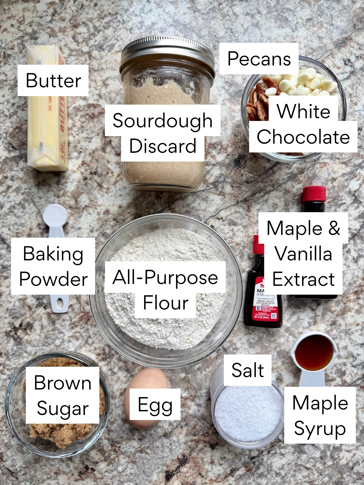 The ingredients to make maple pecan sourdough blondies. Each ingredient is labeled with text. They include butter, sourdough discard, pecans, white chocolate, baking powder, all-purpose flour, maple and vanilla extract, brown sugar, egg, salt, and maple syrup.