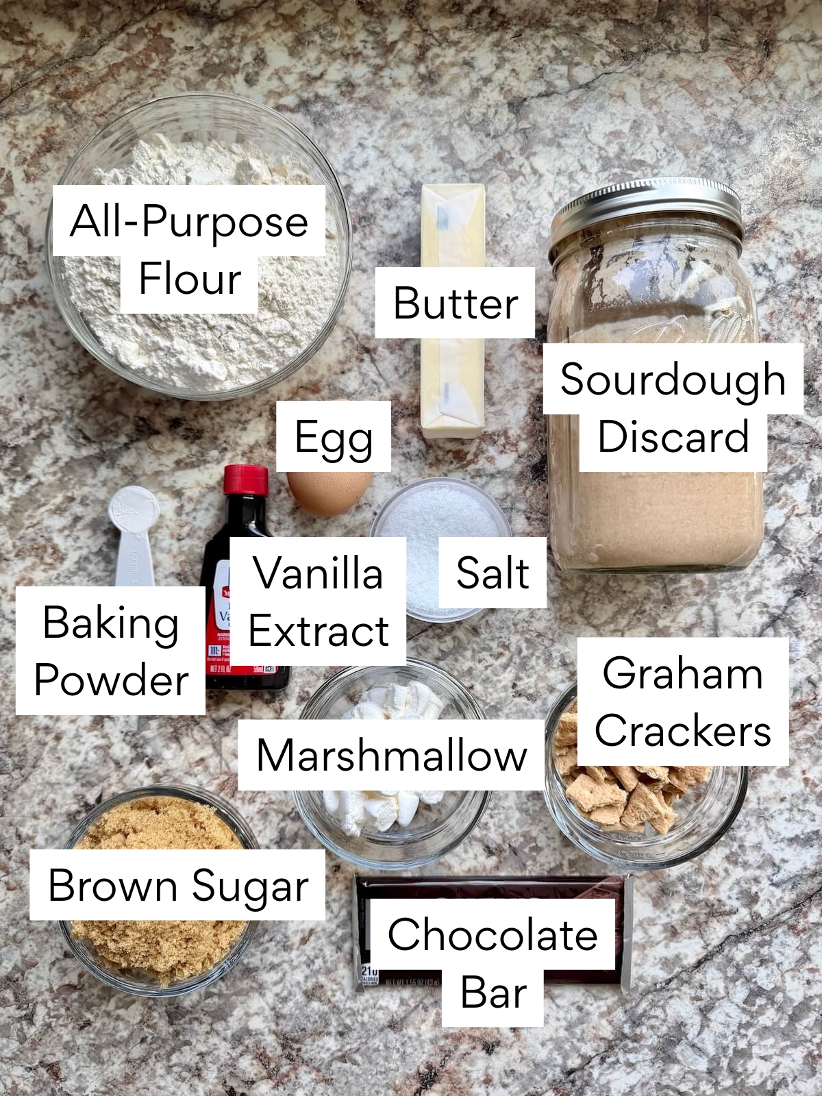 The ingredients to make s'more sourdough blondies. Each ingredient is labeled with text. They include all-purpose flour, butter, sourdough discard, egg, baking powder, vanilla extract, salt, brown sugar, marshmallow, graham crackers, and chocolate bar.