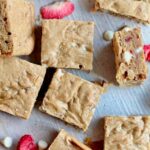 Several strawberry sourdough blondies on a piece of parchment paper with white chocolate chips and freeze-dried strawberries scattered around them.