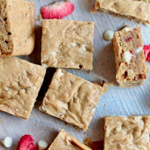 Several strawberry sourdough blondies on a piece of parchment paper with white chocolate chips and freeze-dried strawberries scattered around them.