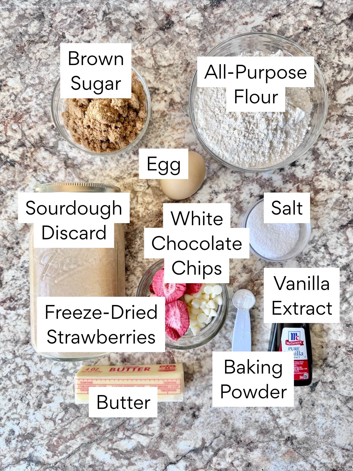 The ingredients to make sourdough strawberry blondies. Each ingredient is labeled with text. They include brown sugar, all-purpose flour, egg, sourdough discard, salt, white chocolate chips, freeze-dried strawberries, butter, vanilla extract, and baking powder.