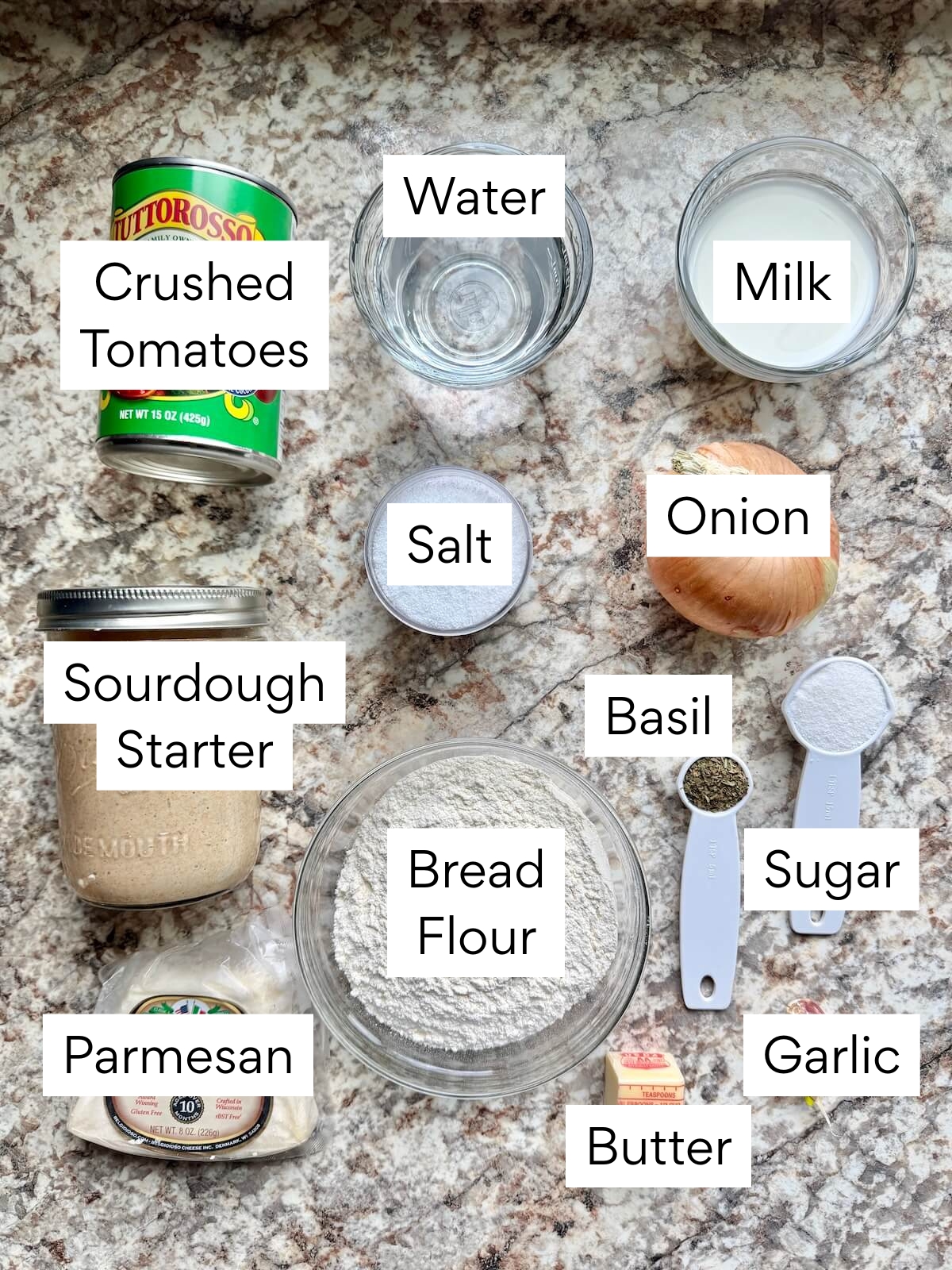 The ingredients to make tomato basil sourdough bread. Each ingredient is labeled with text. They include crushed tomatoes, water, milk, sourdough starter, salt, onion, bread flour, basil, sugar, parmesan, butter, and garlic.