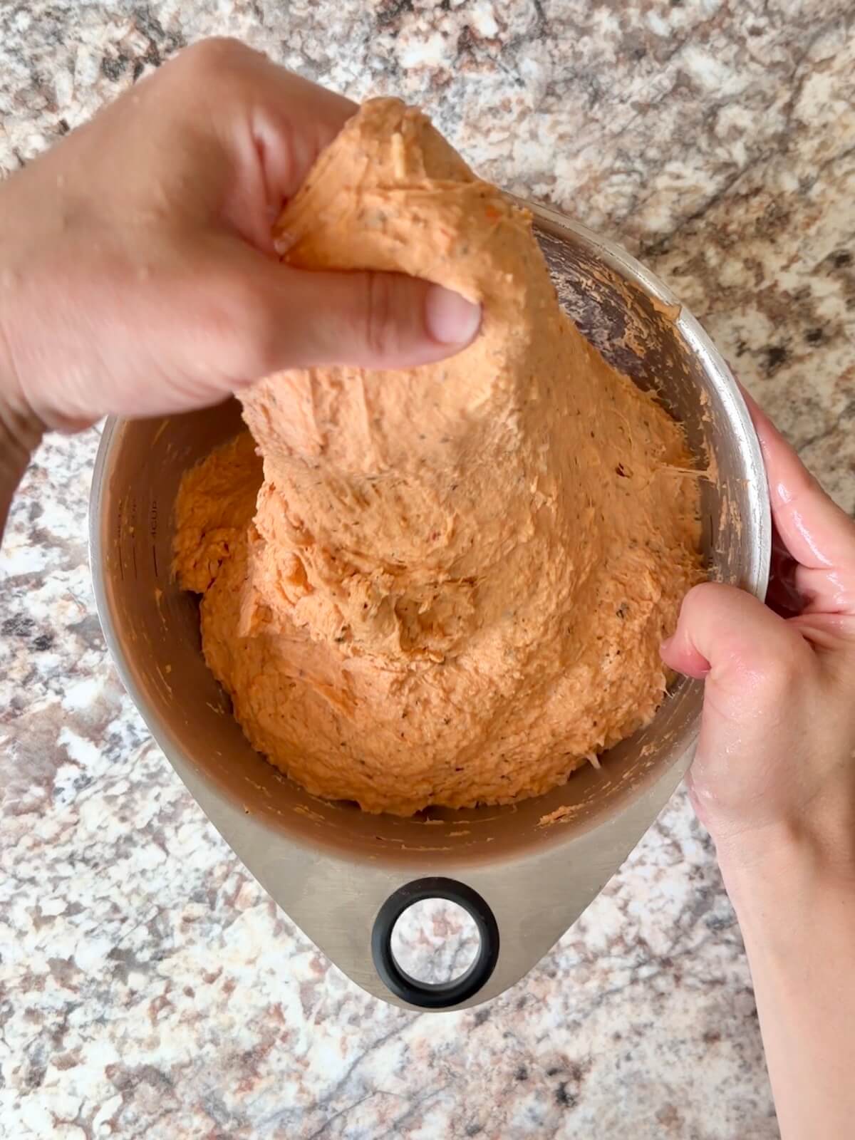 Two hands stretching the tomato basil sourdough bread dough up out of the mixing bowl.