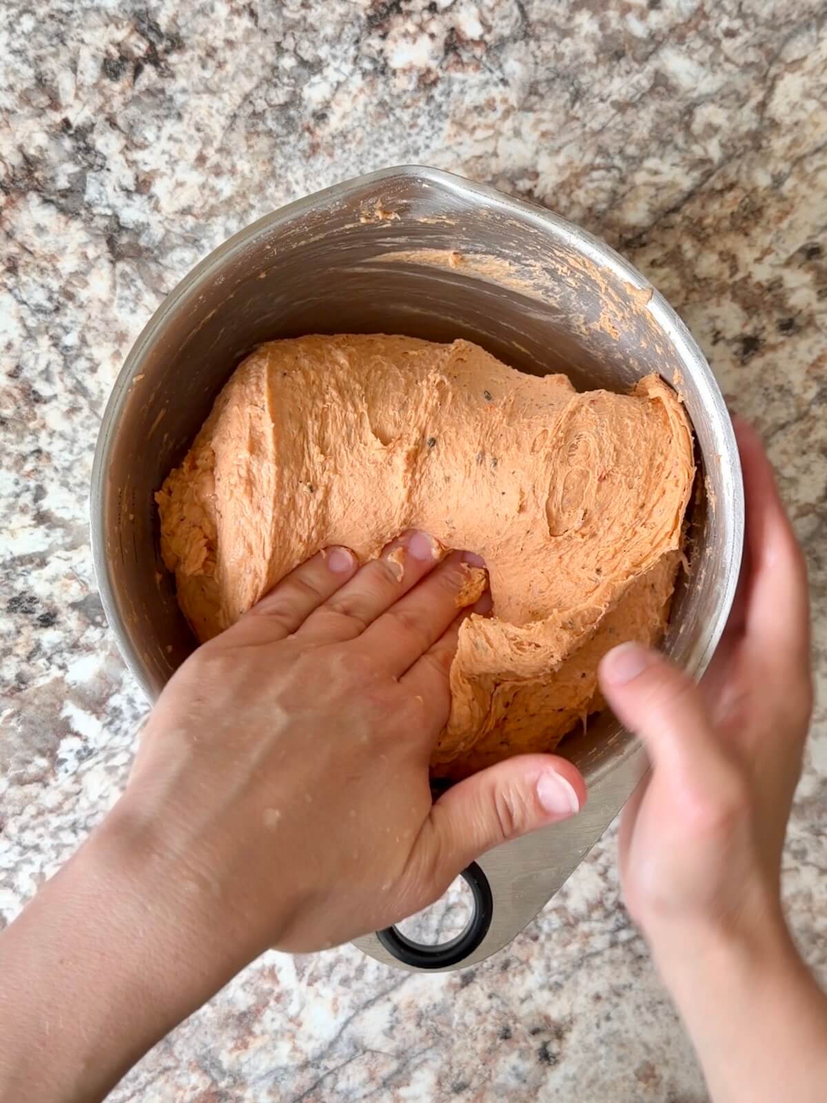 Two hands folding the tomato soup sourdough bread dough over itself in a mixing bowl.