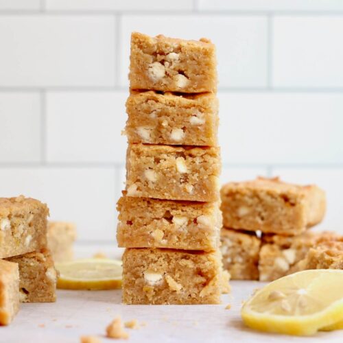 A stack of five lemon sourdough blondies with more on the counter surrounding the stack. There are several lemon slices also on the counter.