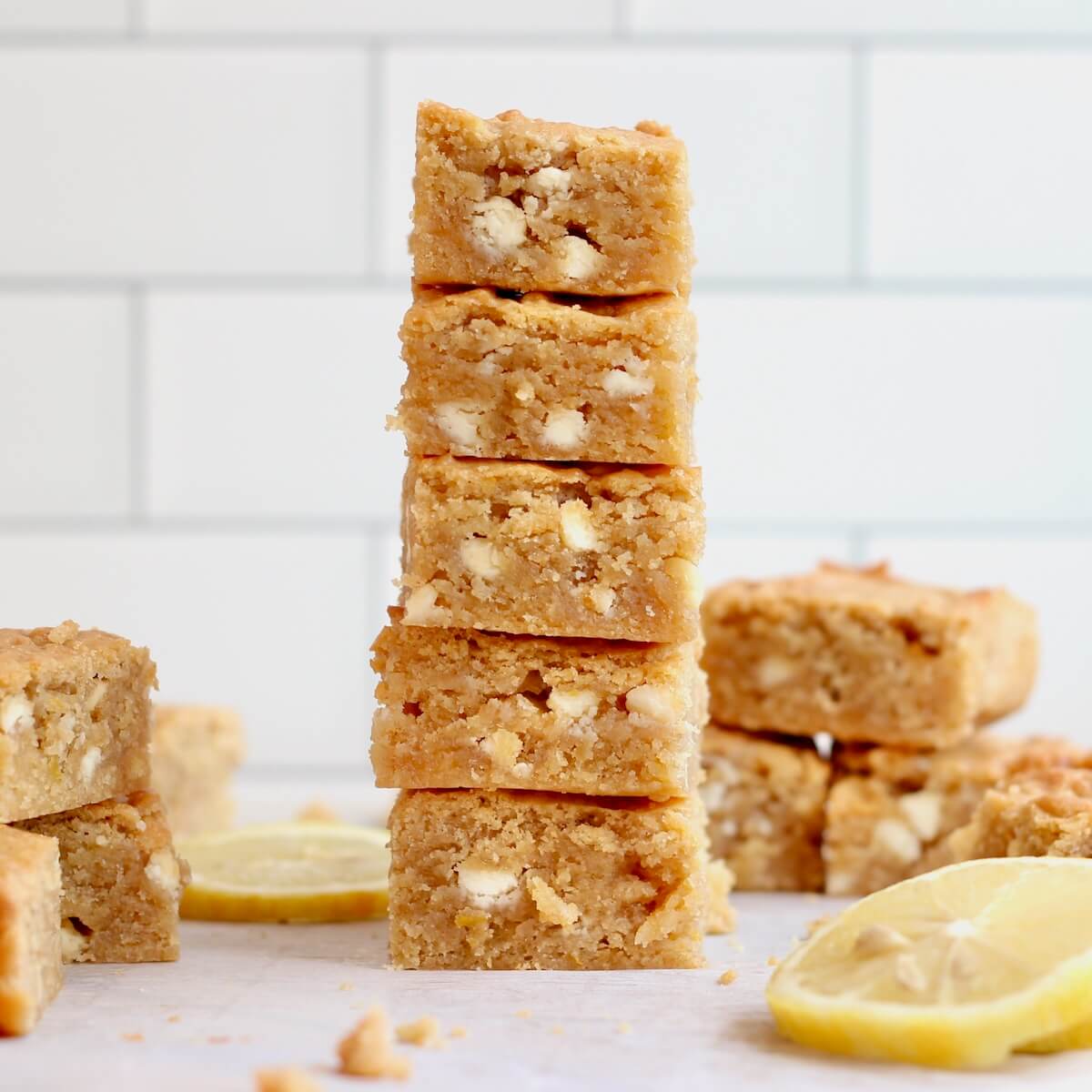 A stack of five lemon sourdough blondies with more on the counter surrounding the stack. There are several lemon slices also on the counter.