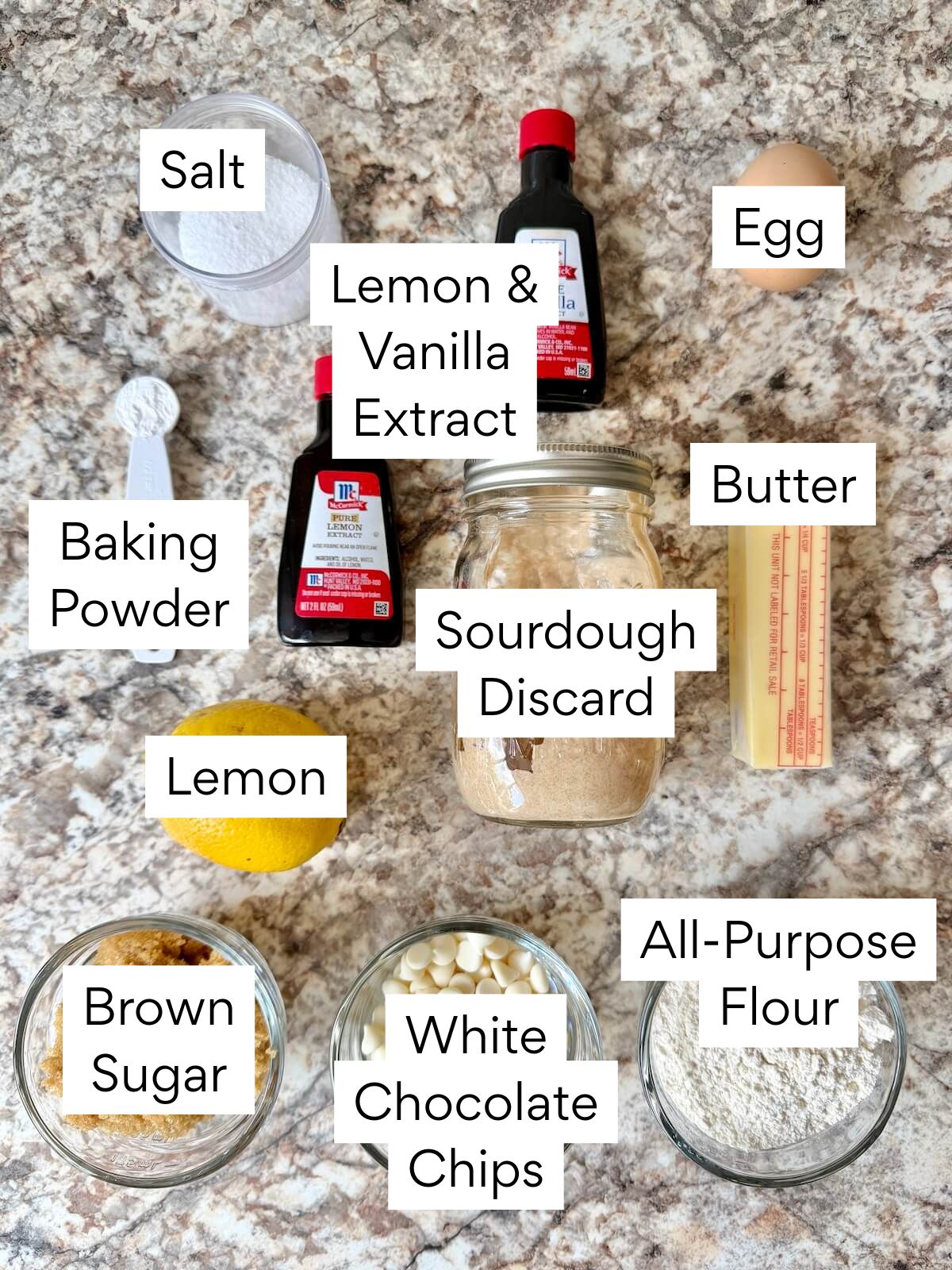 The ingredients to make sourdough lemon blondies. Each ingredient is labeled with text. They include salt, lemon and vanilla extract, egg, baking powder, sourdough discard, butter, lemon, brown sugar, white chocolate chips, and all-purpose flour.