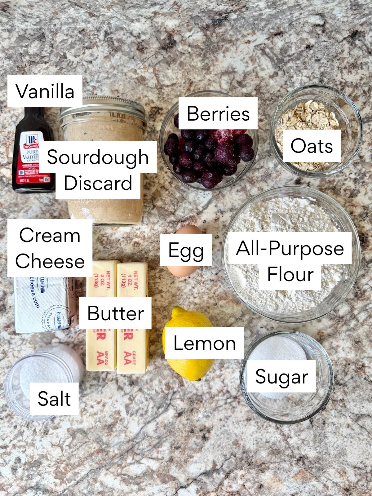 The ingredients to make sourdough raspberry crumble bars. Each ingredient is labeled with text. They include vanilla, sourdough discard, berries, oats, cream cheese, egg, butter, all-purpose flour, salt, lemon, and sugar.
