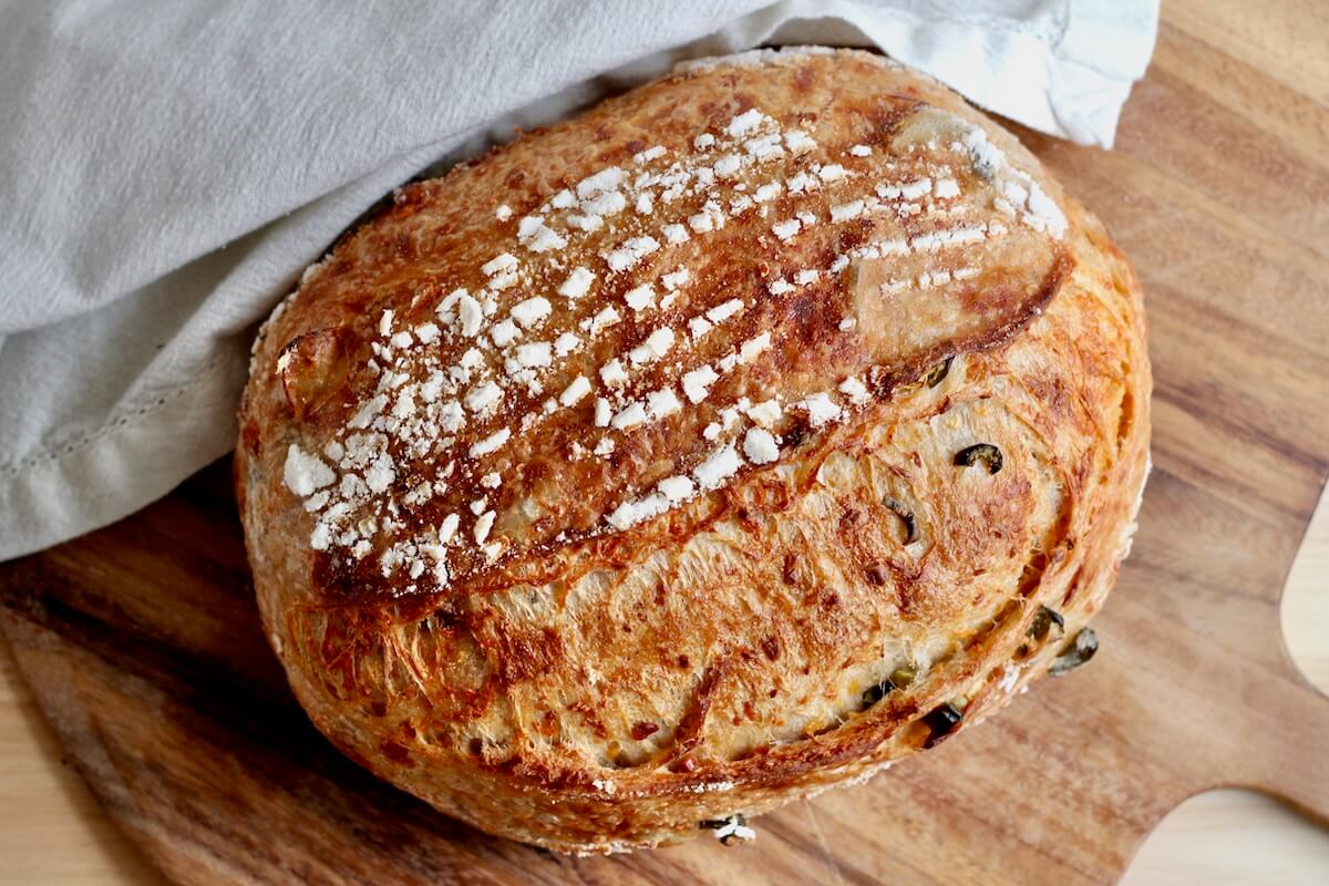 A whole loaf of jalapeƱo cheddar sourdough bread on a wooden cutting board next to a white cloth napkin.
