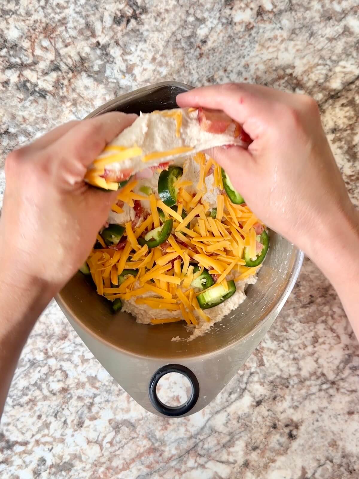 JalapeƱos, shredded cheddar, and bacon being folded into the dough in a bowl.