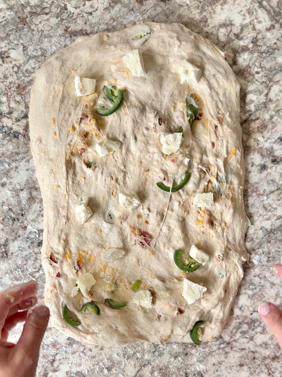 The fermented dough stretched into a rectangle on a floured counter. Chunks of cream cheese are placed all over the dough.