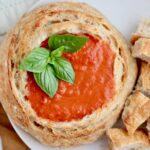 A sourdough bread bowl filled with tomato soup on a small plate with chunks of sourdough bread on the plate to the right. The soup is garnished with fresh basil.