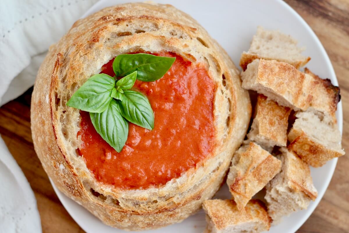 A sourdough bread bowl filled with tomato soup on a small plate with chunks of sourdough bread on the plate to the right. The soup is garnished with fresh basil.