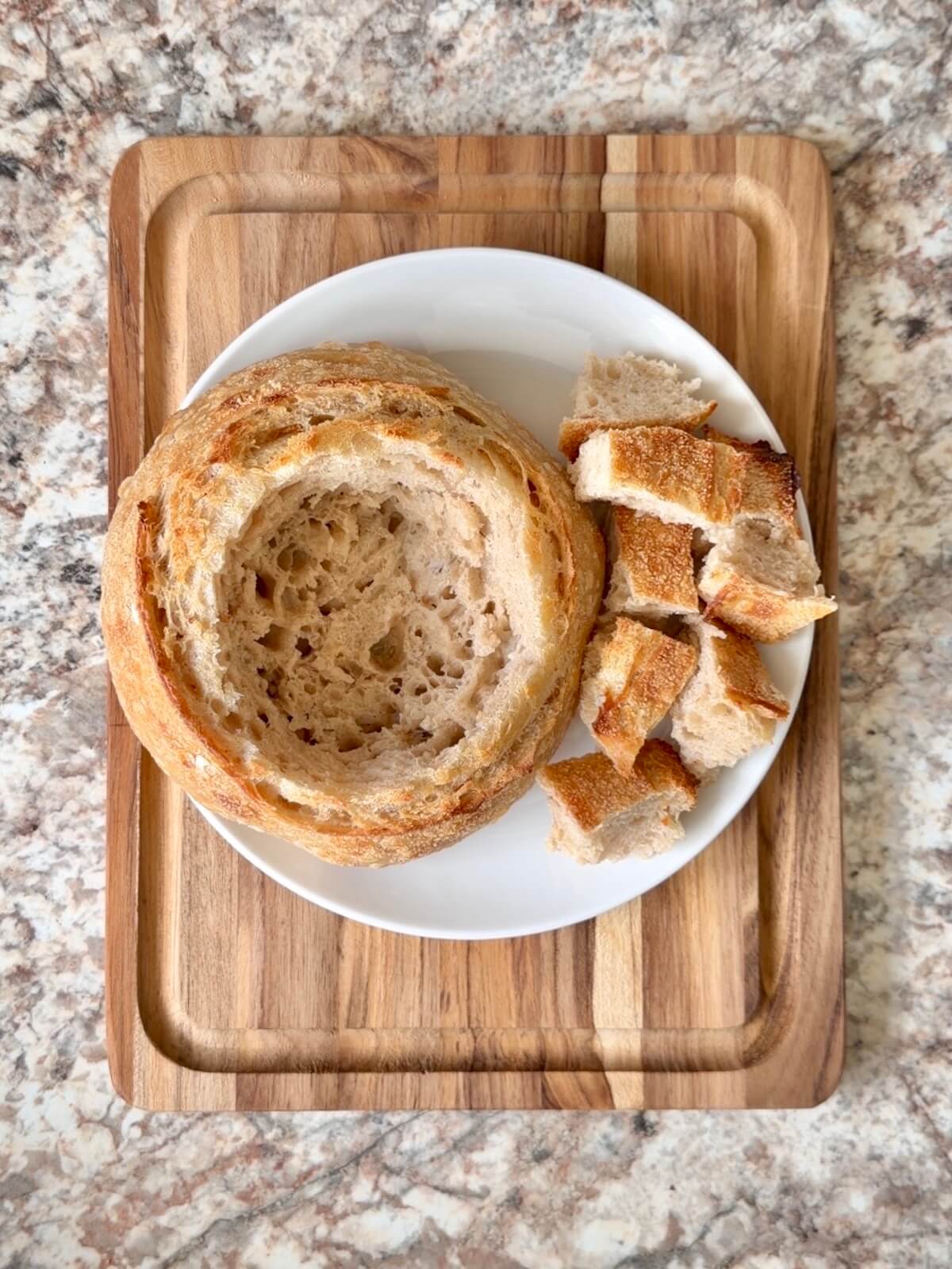 One sourdough bread bowl hollowed out on a small plate with the bread chunks sitting on the plate to the right.