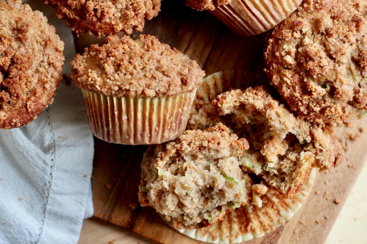 Several sourdough zucchini muffins with cinnamon streusel topping on a wooden counter. One of the muffins is removed from the liner and split in half to show the crumb.