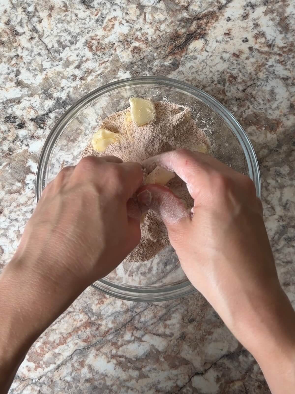 Two hands pinching butter into flour, sugar, and cinnamon in a bowl.