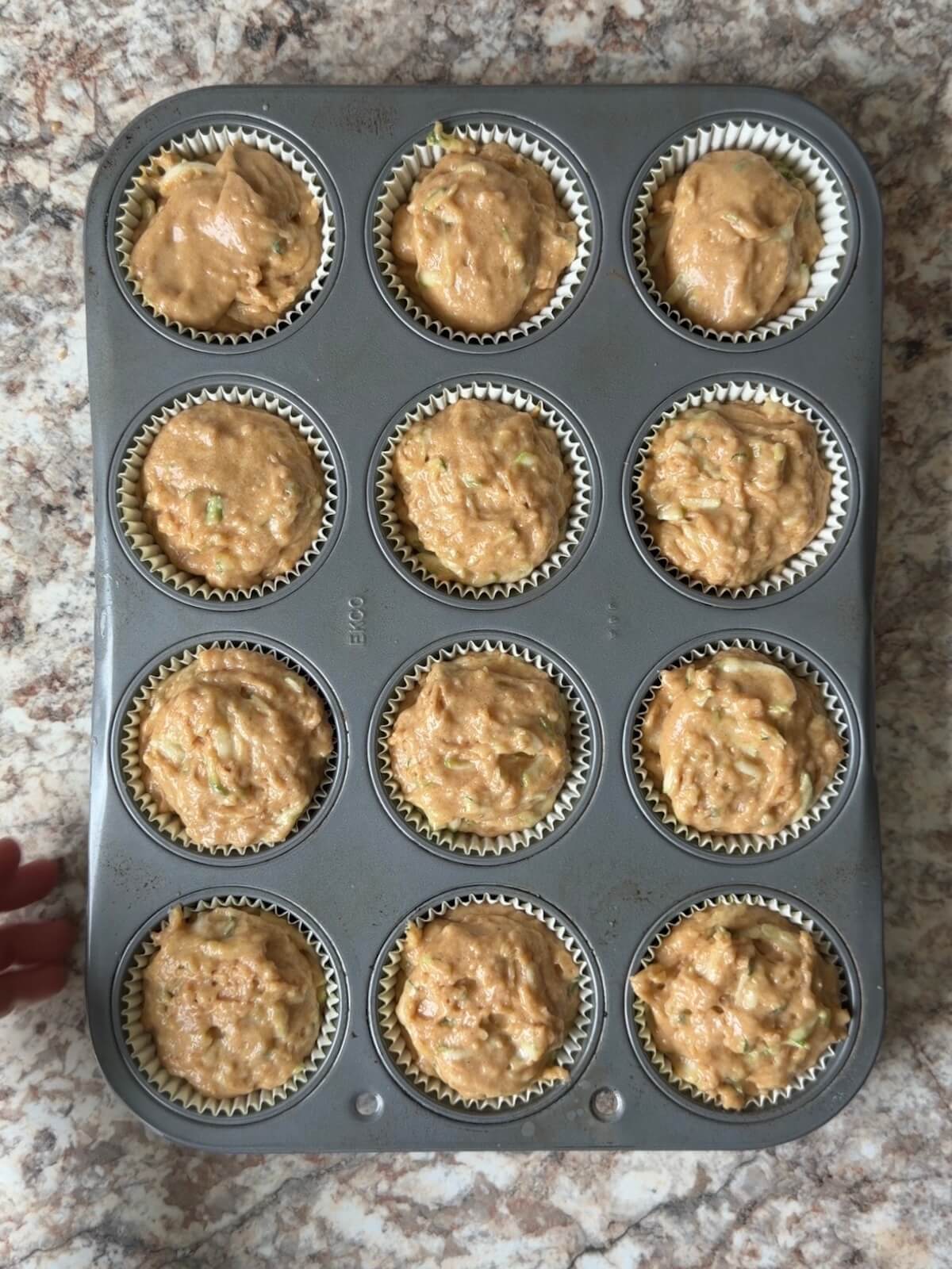A standard 12-cup muffin pan with paper liners filled with sourdough discard zucchini muffin batter.