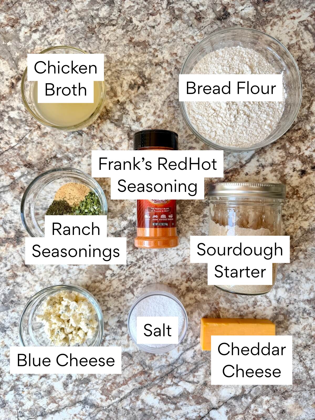 The ingredients to make buffalo chicken dip sourdough bread. Each ingredient is labeled with text. They include chicken broth, bread flour, Frank's RedHot Seasoning, ranch seasonings, sourdough starter, blue cheese, salt, and cheddar cheese.