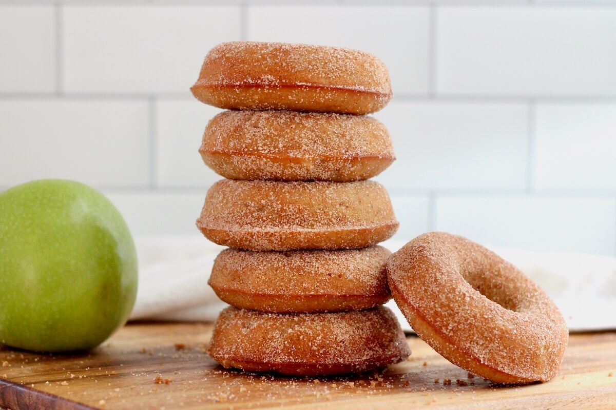 A stack of five sourdough apple cider donuts with one more donut leaning against them and a whole green apple out of focus in the background.