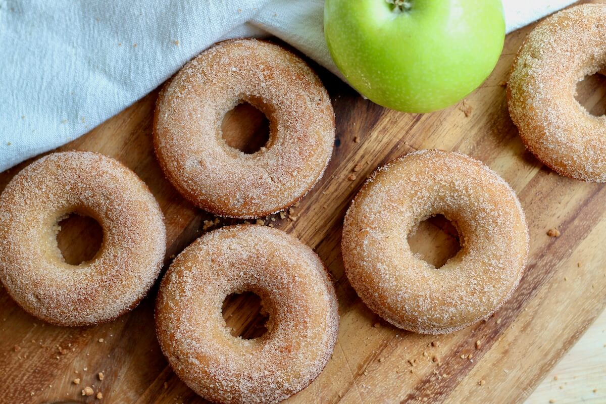 Five sourdough apple cider donuts on a wooden cutting board with a whole green apple.