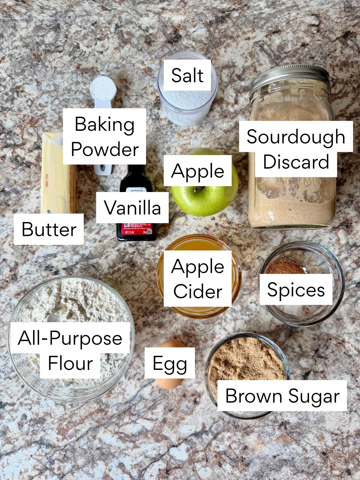 The ingredients to make sourdough apple cider donuts. Each ingredient is labeled with text. They include butter, baking powder, vanilla, salt, apple, sourdough discard, all-purpose flour, apple cider, spices, egg, and brown sugar.
